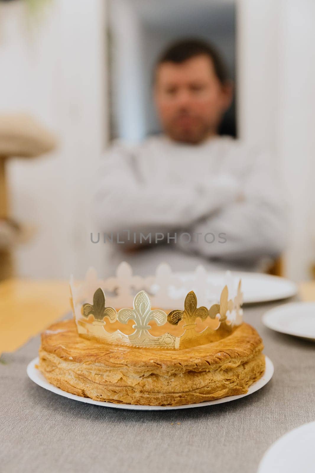 One delicious puff royal galette with a golden paper crown lies in a plate on a wooden table, against the background of a blurred man, side view close-up.