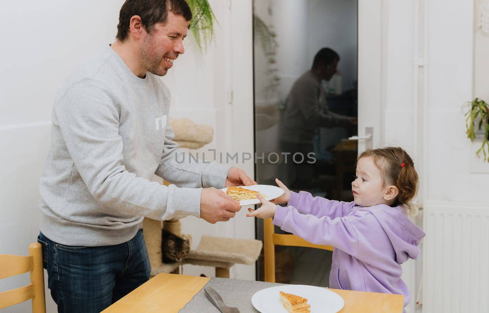 One little Caucasian girl takes a plate with a piece of royal biscuit from the hands of a happy and smiling father standing at the table in the kitchen near the glass door with access to the backyard, close-up side view.