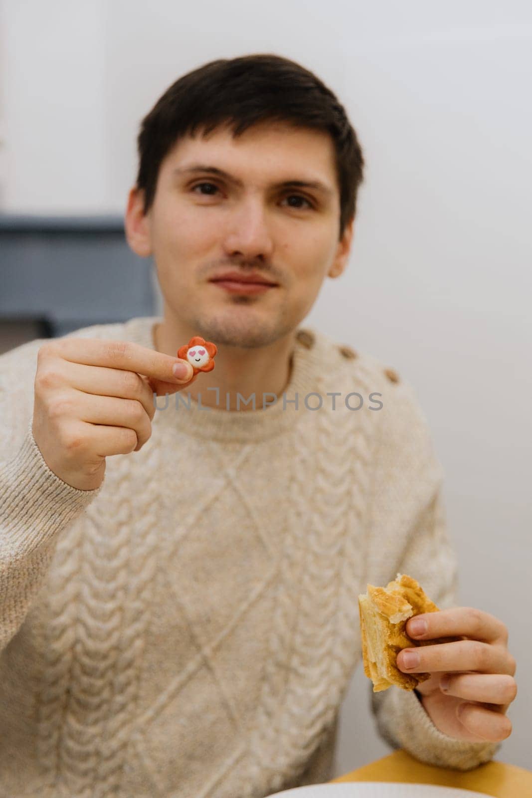 One young handsome Caucasian guy with a smile shows a surprise toy from an eaten piece of royal galette, sitting at a table in the kitchen, close-up side view.