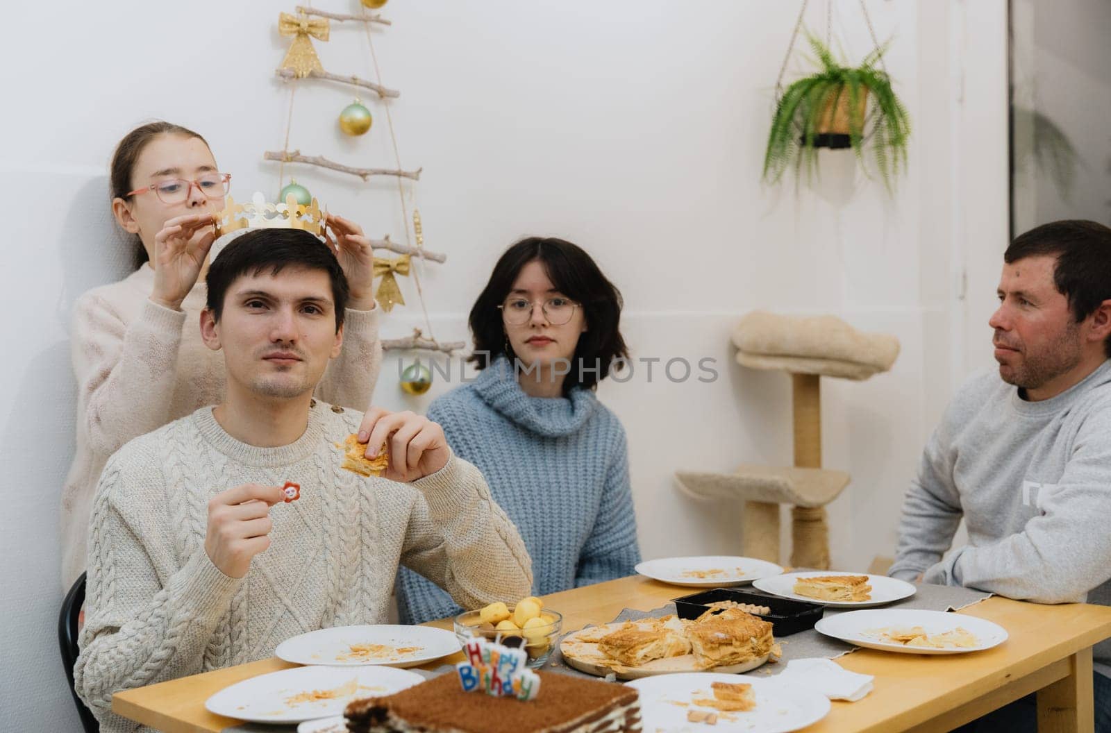 One beautiful Caucasian girl with glasses solemnly puts on a golden paper crown to an adult guy who ate a royal galette and found it and with a smile shows a surprise gift, sitting at the table in the kitchen with his family, close-up side view.