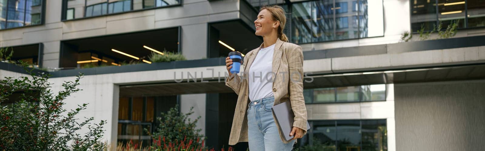 Smiling female freelancer with laptop and take away coffee during break time near modern building by Yaroslav_astakhov