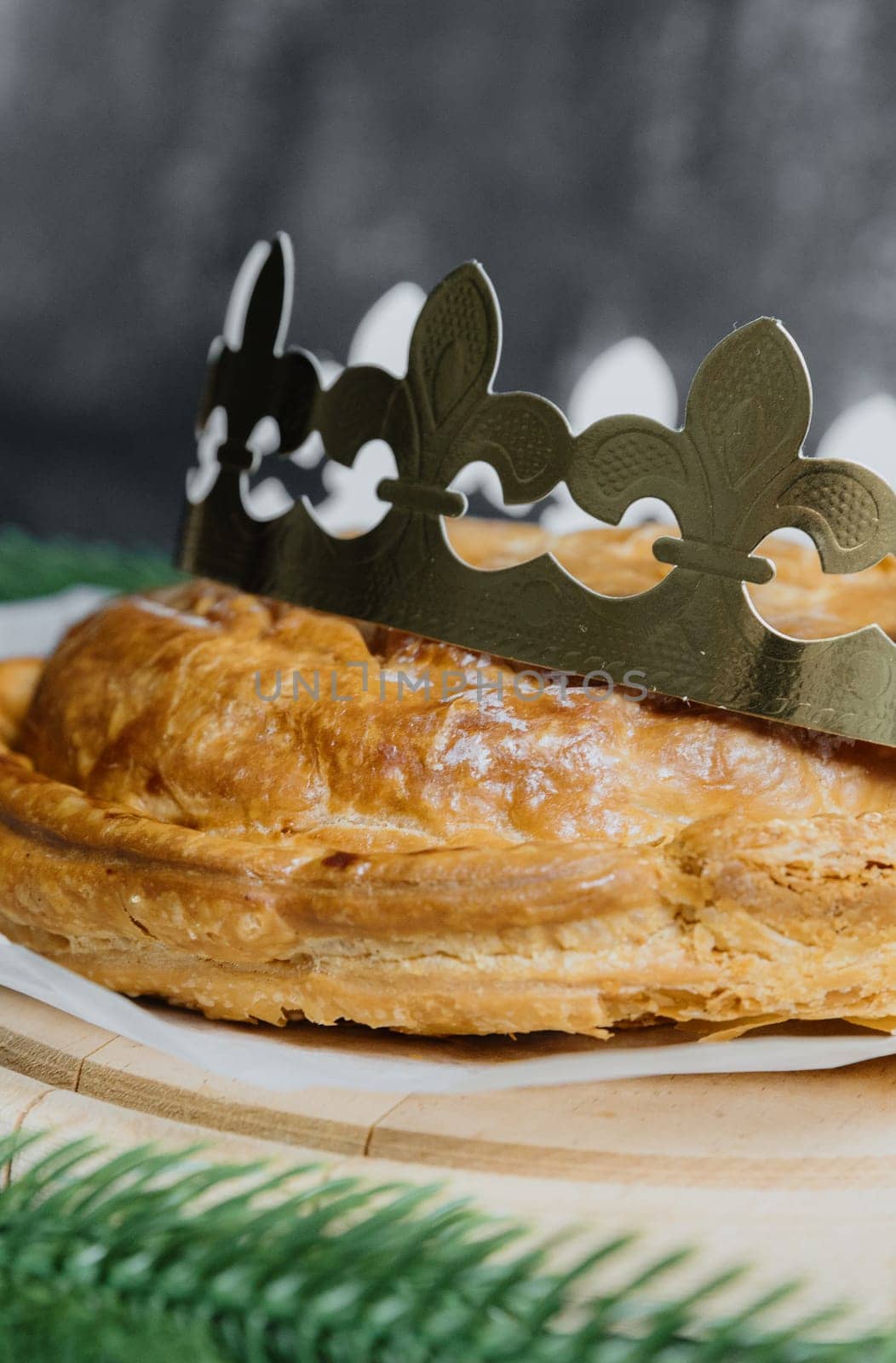 One whole freshly baked golden crown king galette on a round wooden cutting board with baking paper stands on the table with a blurred dark gray background, side view close up with depth of field.