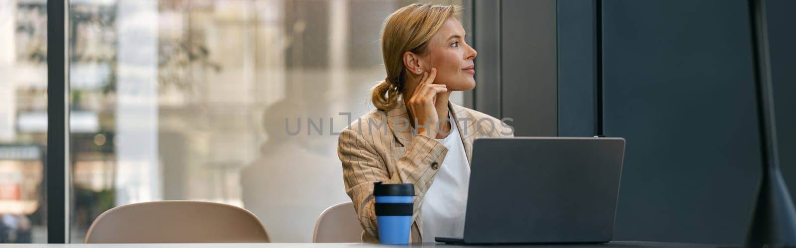 Stylish business woman working on laptop sitting the desk on office background and looks away by Yaroslav_astakhov