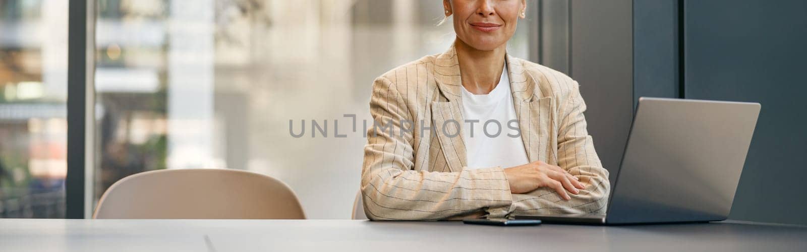 Focused business woman working on laptop sitting the desk on office background and looks camera by Yaroslav_astakhov