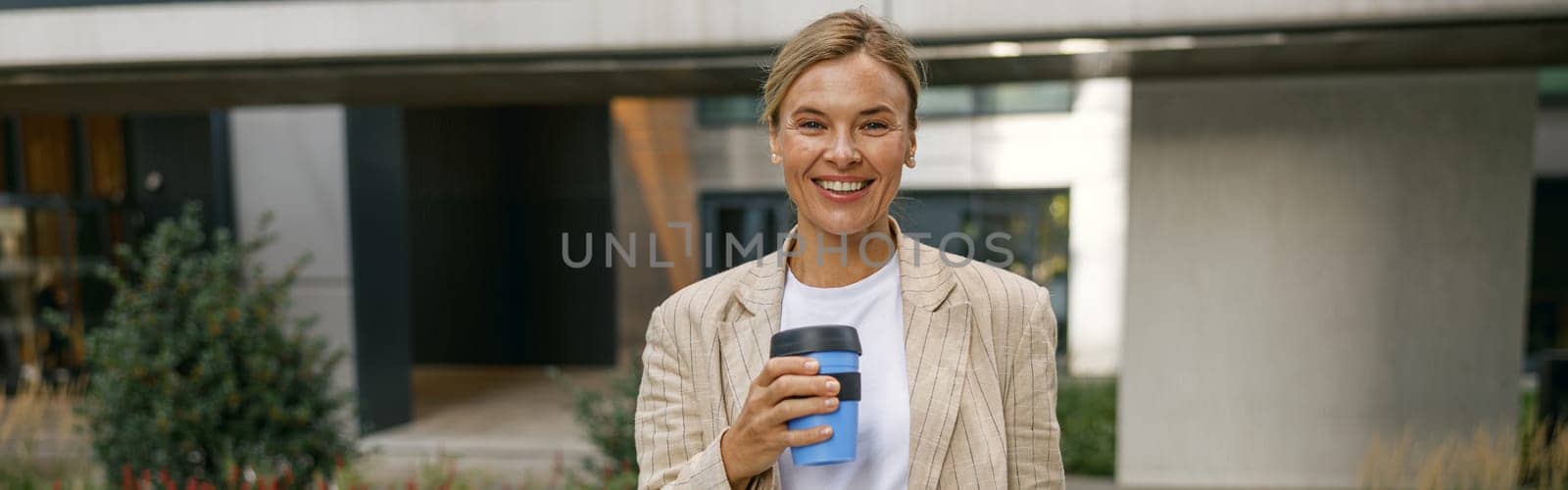 Business woman going to work while drinking coffee on office building background and looks camera by Yaroslav_astakhov
