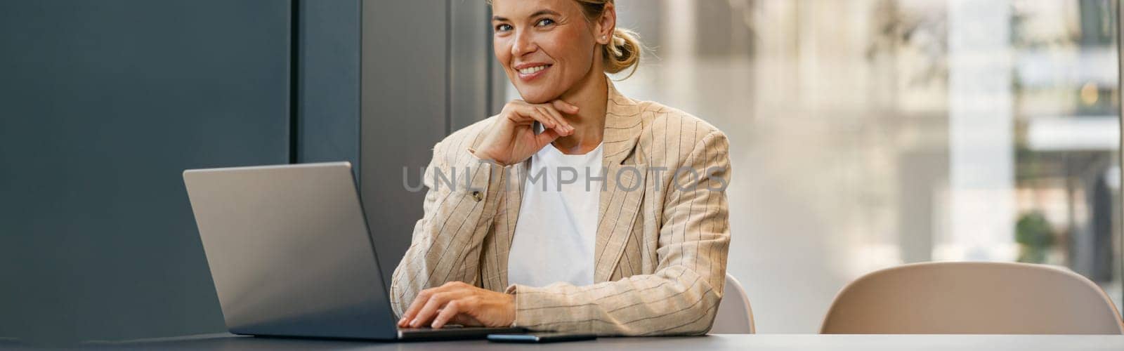 Smiling woman manager working on laptop sitting the desk on office background and looks camera by Yaroslav_astakhov