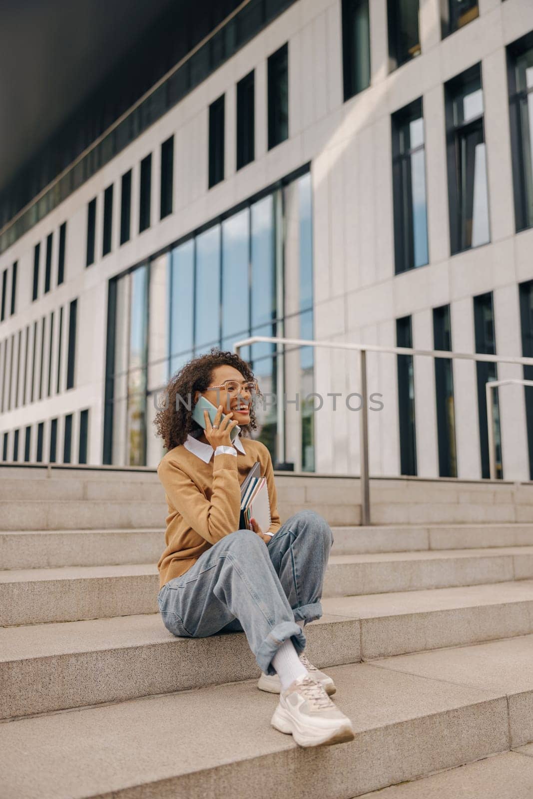 Smiling female freelancer talking phone with client while sitting on building background by Yaroslav_astakhov