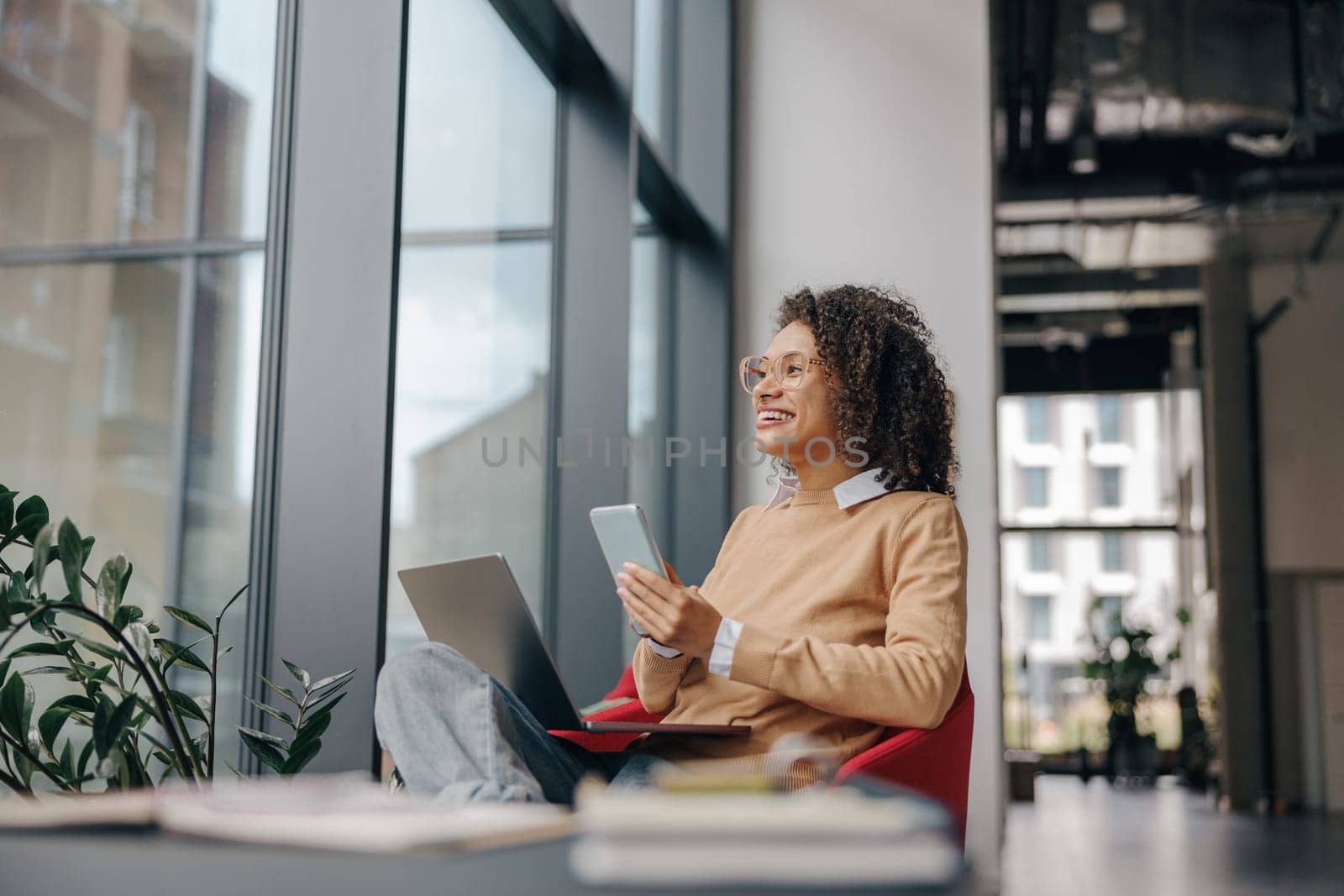 Stylish business woman is use phone while working on laptop in modern office near window