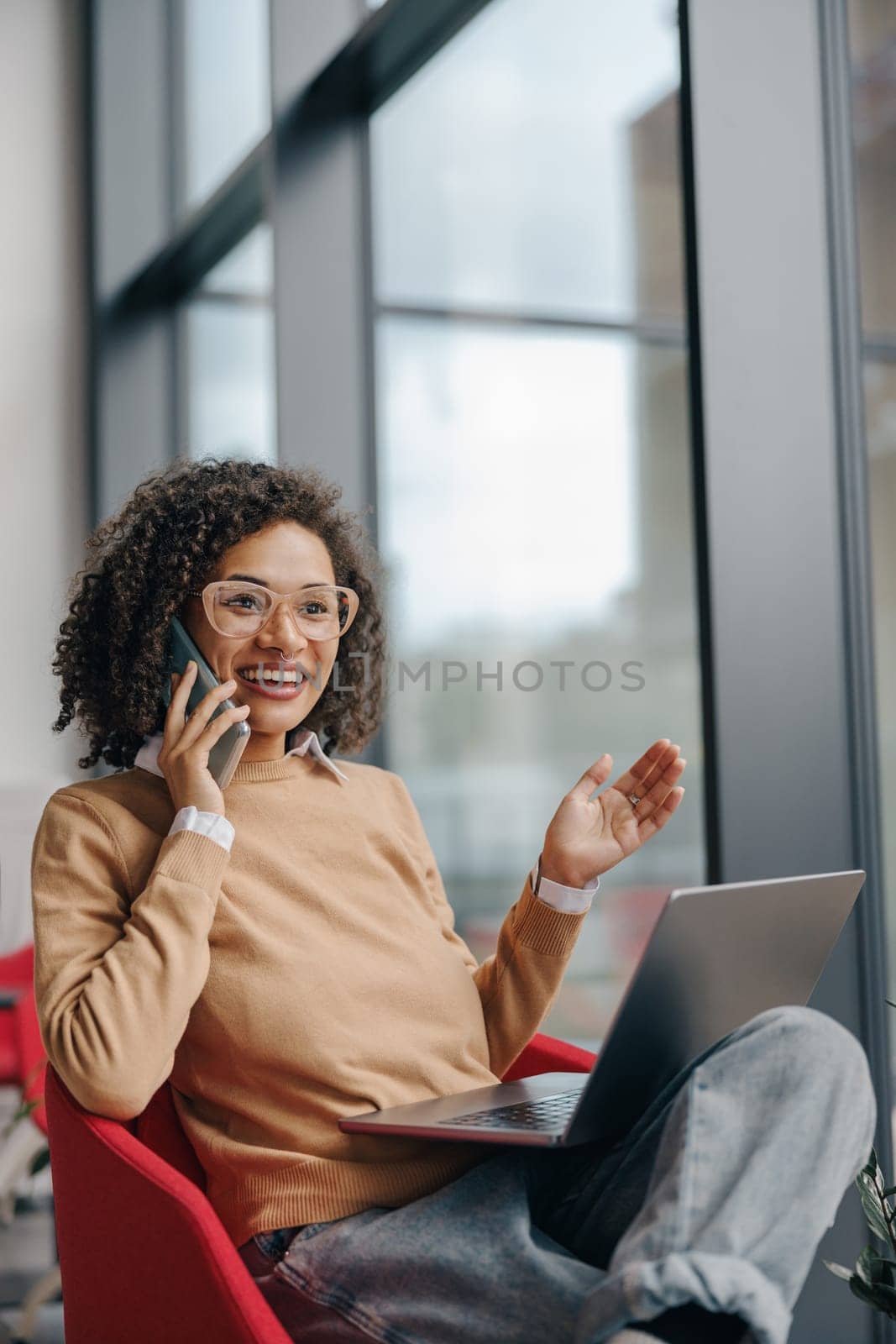 Stylish business woman is talking phone while working on laptop in modern office near window