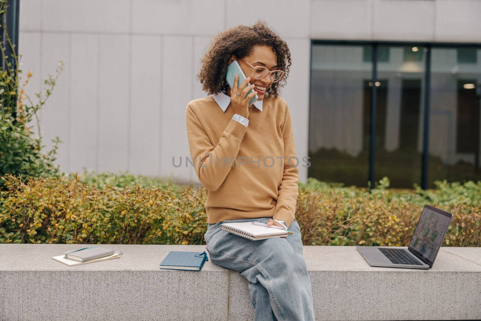 Smiling female freelancer talking phone and making notes while sitting on building background