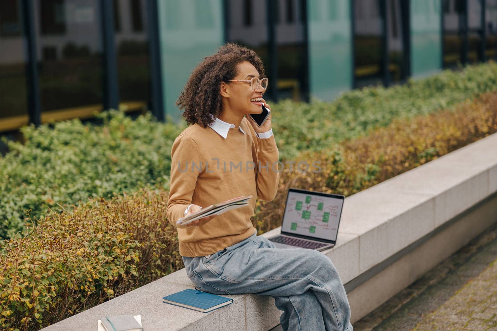 Smiling female freelancer talking phone with client while sitting on building background