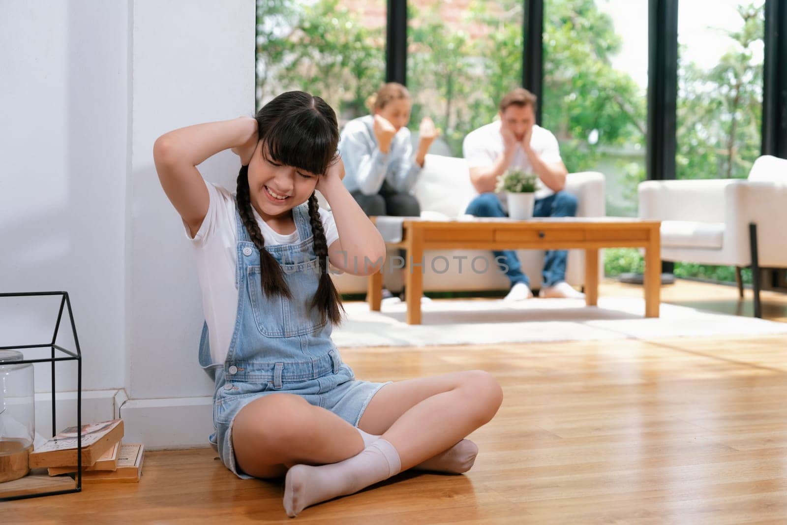 Stressed and unhappy girl huddle in corner, cover her ears with painful expression while her parent arguing in background. Domestic violence and traumatic childhood develop to depression. Synchronos