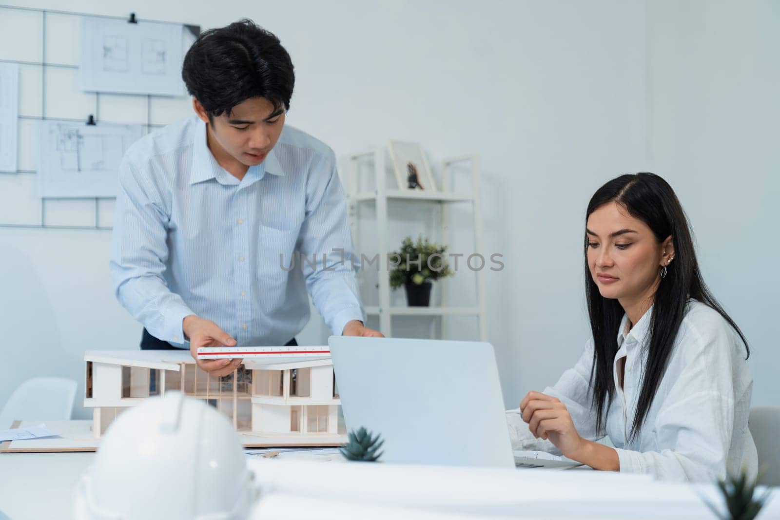 Professional male asian architect using ruler to measure house model length while young beautiful caucasian colleague using laptop to analyzed data on meeting table with house model. Immaculate.