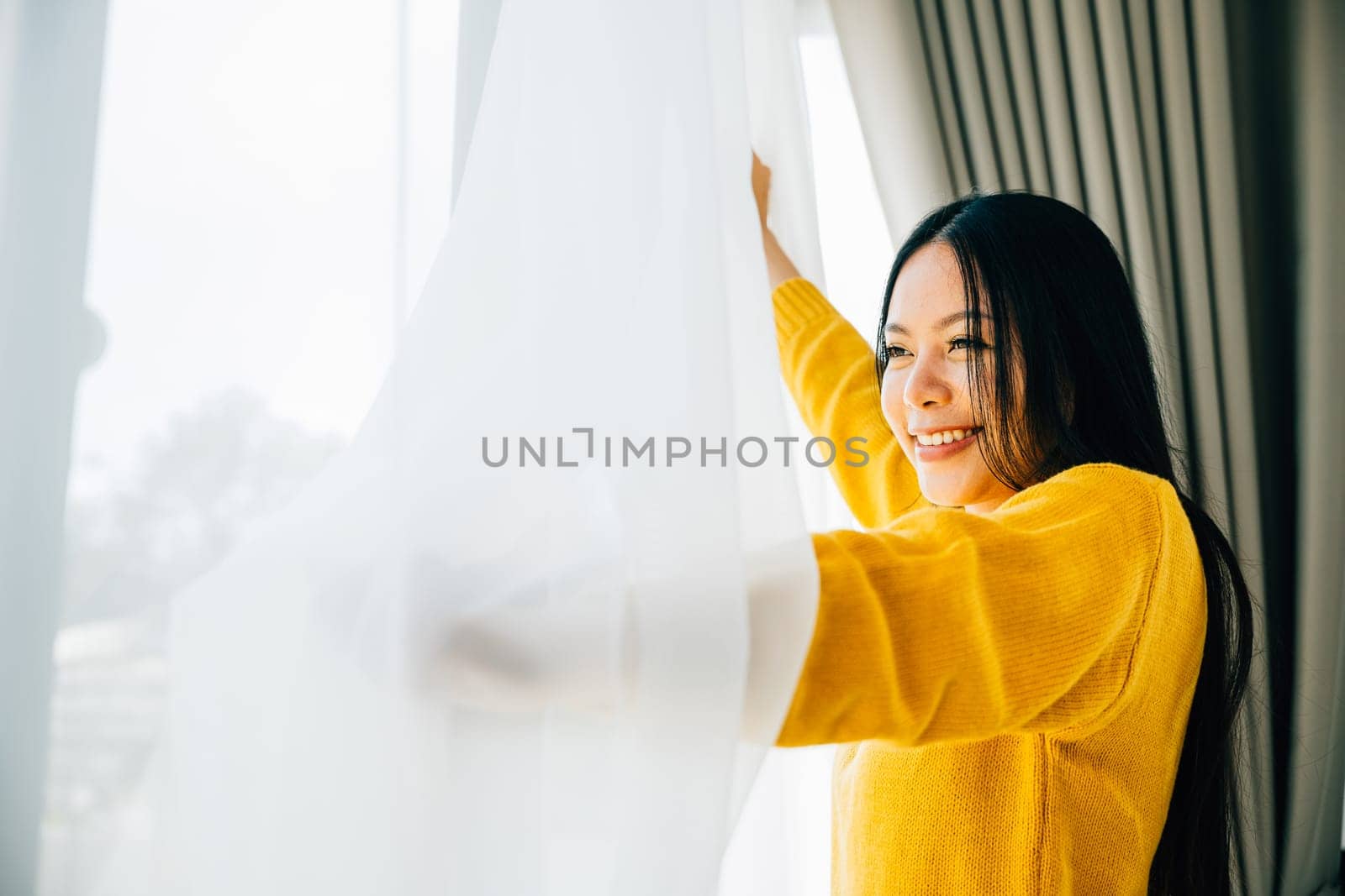 A young woman opens curtains in the early morning smiles at the view feeling relaxed by Sorapop