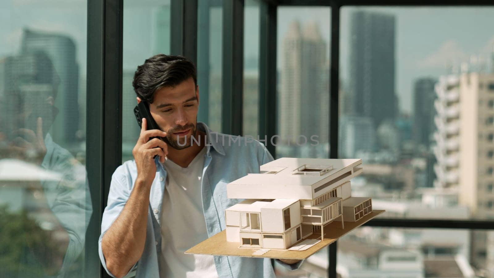 Smart caucasian engineer standing and holding house model while phone calling to manager. Professional manager inspect at architectural model while standing near window. Design concept. Tracery