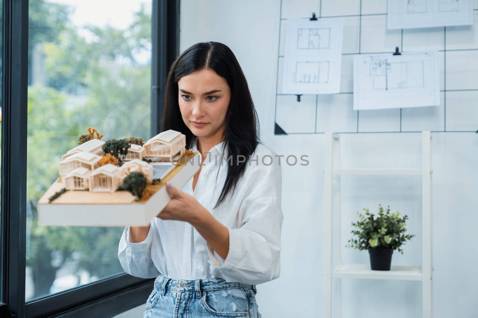 Close up of skilled young beautiful caucasian architect lift house model to check and focus on inspecting mistake point at modern office with blueprint. Creative design and living concept. Immaculate.