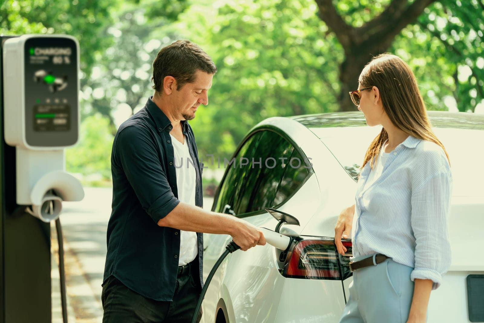 Lovely young couple wearing sun glasses recharging battery for electric car during road trip travel EV car in natural forest or national park. Eco friendly travel during vacation and holiday. Exalt