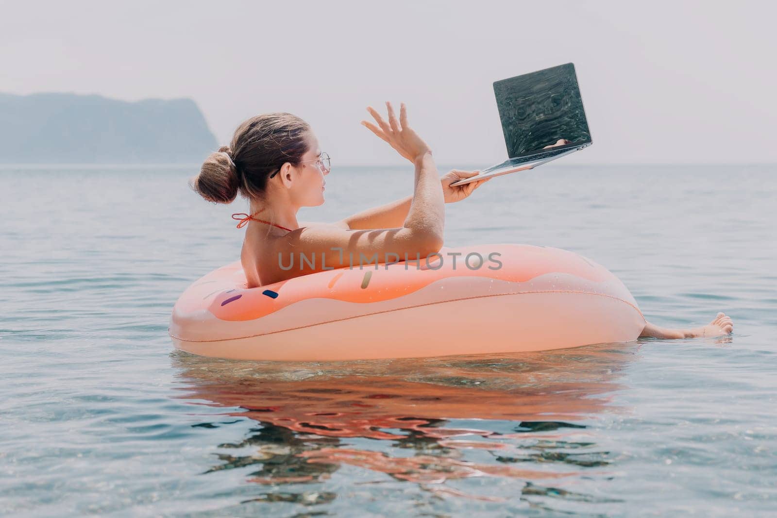 Woman freelancer works on laptop swimming in sea on pink inflatable ring. Happy tourist floating on inflatable donut and working on laptop computer in calm ocean. Freelance, remote working anywhere by panophotograph