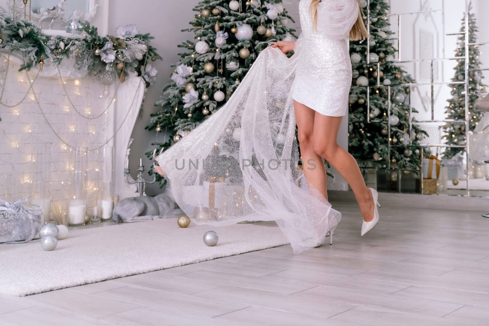 Women's feet christmas tree. Cropped photo of beautiful elegant female legs in white high-heeled shoes and a white short dress, a woman stands against the background of a Christmas tree with gifts.