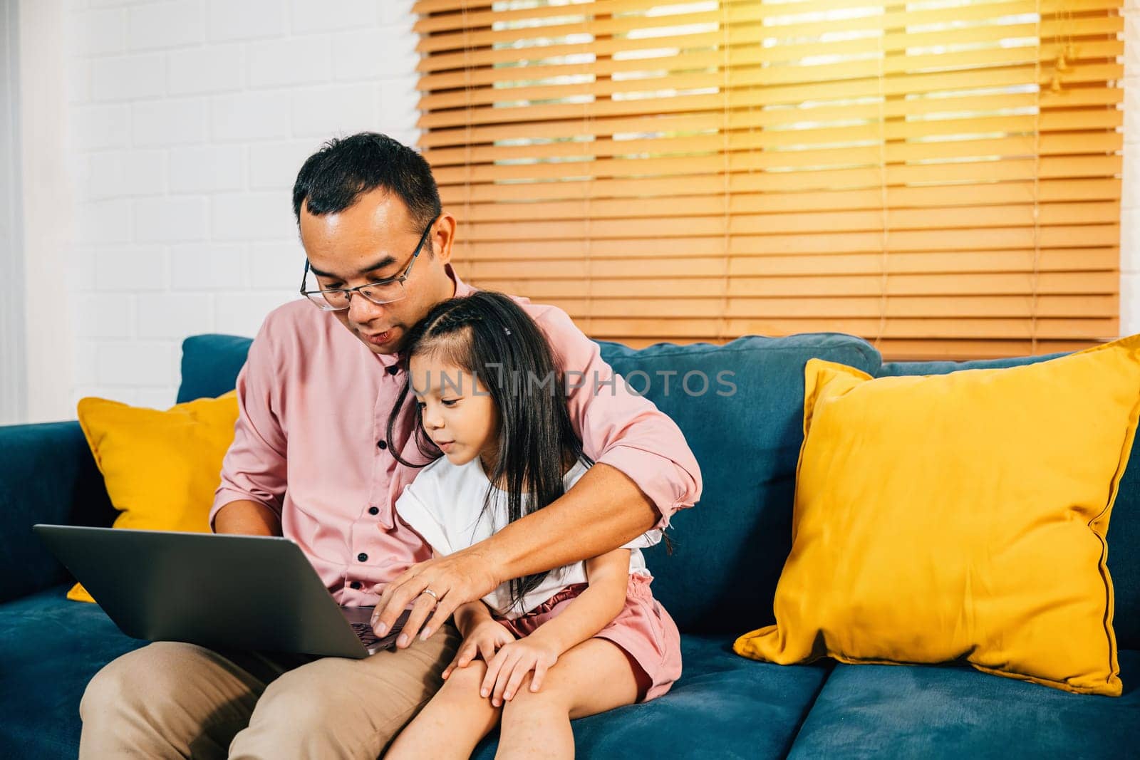 A smiling Asian father works from home on his laptop as his daughter learns on a computer for e-learning by Sorapop