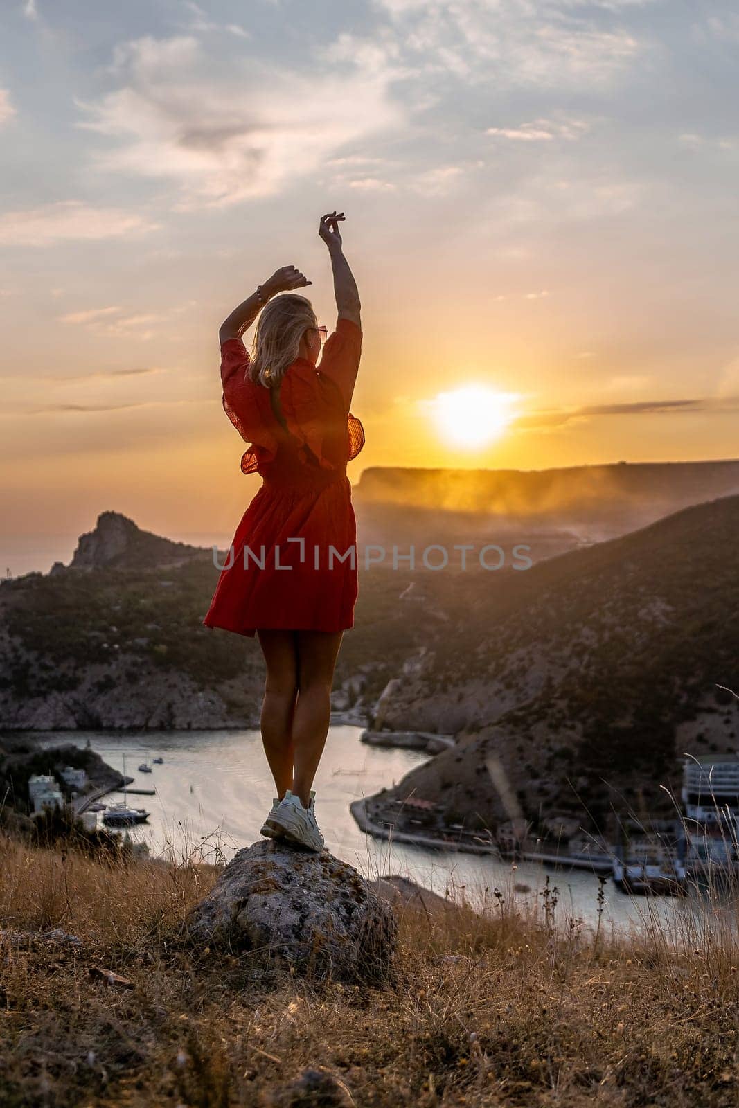 Happy woman standing with her back on the sunset in nature in summer with open hands posing with mountains on sunset, silhouette. Woman in the mountains red dress, eco friendly, summer landscape active rest by Matiunina
