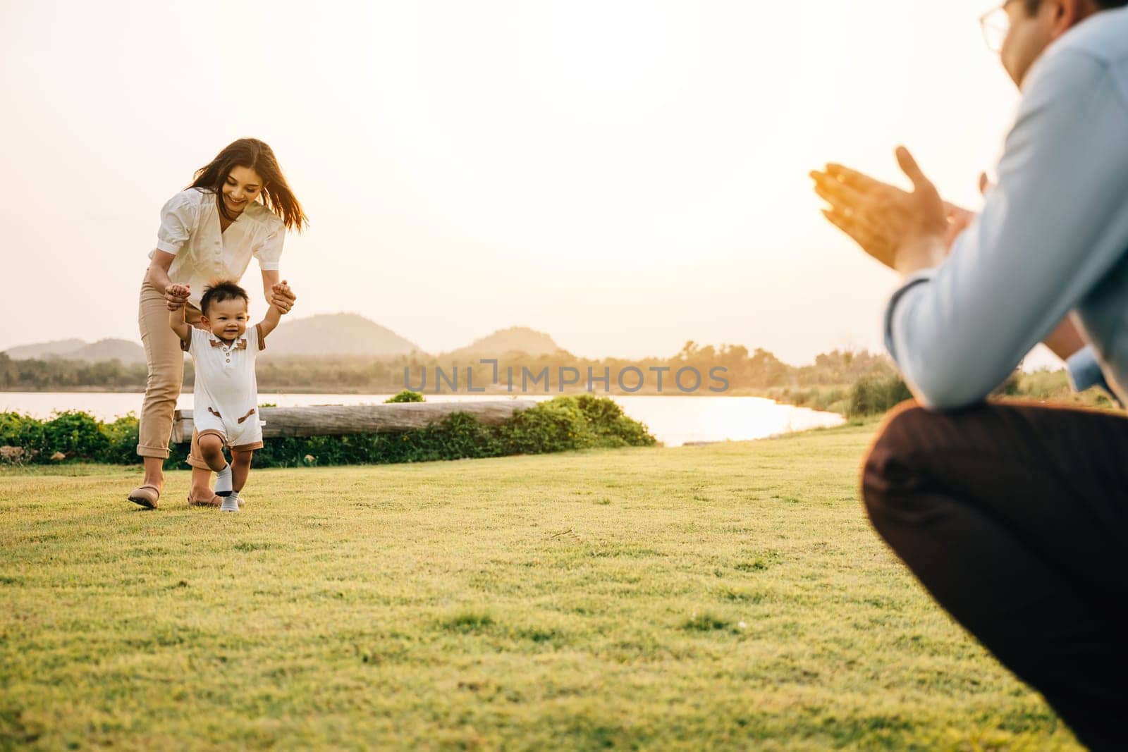 A beautiful Asian family enjoying a sunny day in the park as they teach their baby to take his first steps. Cute toddler little child family concept