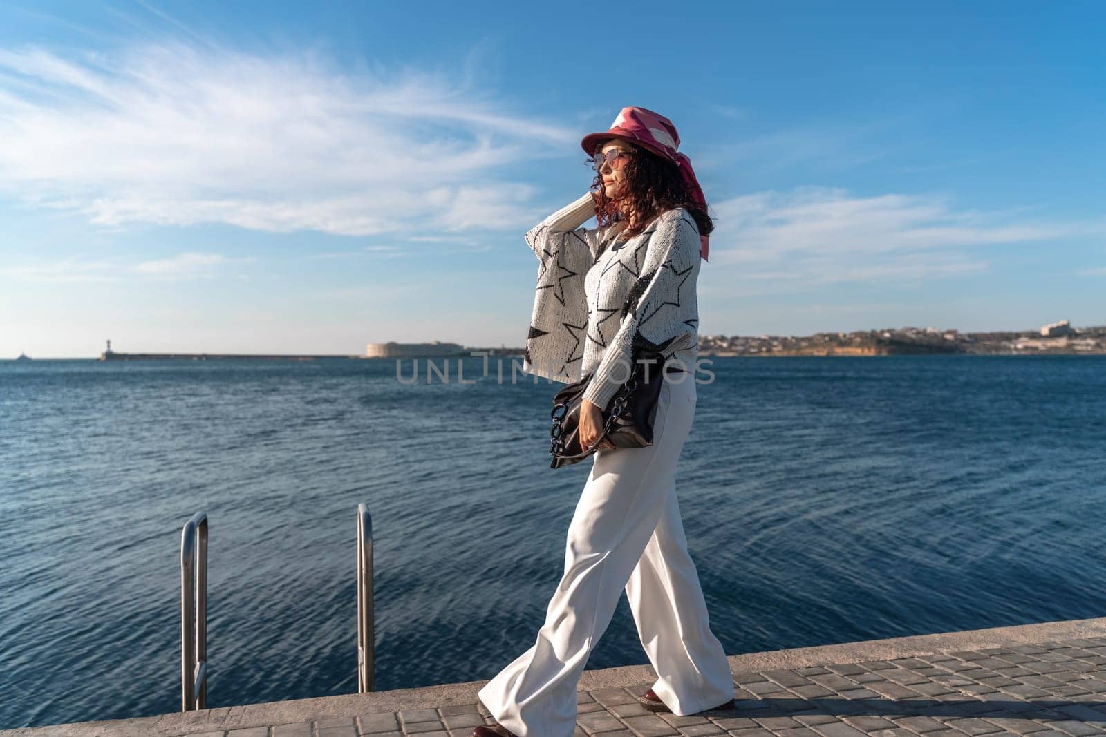 Stylish seashore woman. Fashionable woman in a hat, white trousers and a light sweater with a black pattern on the background of the sea. by Matiunina