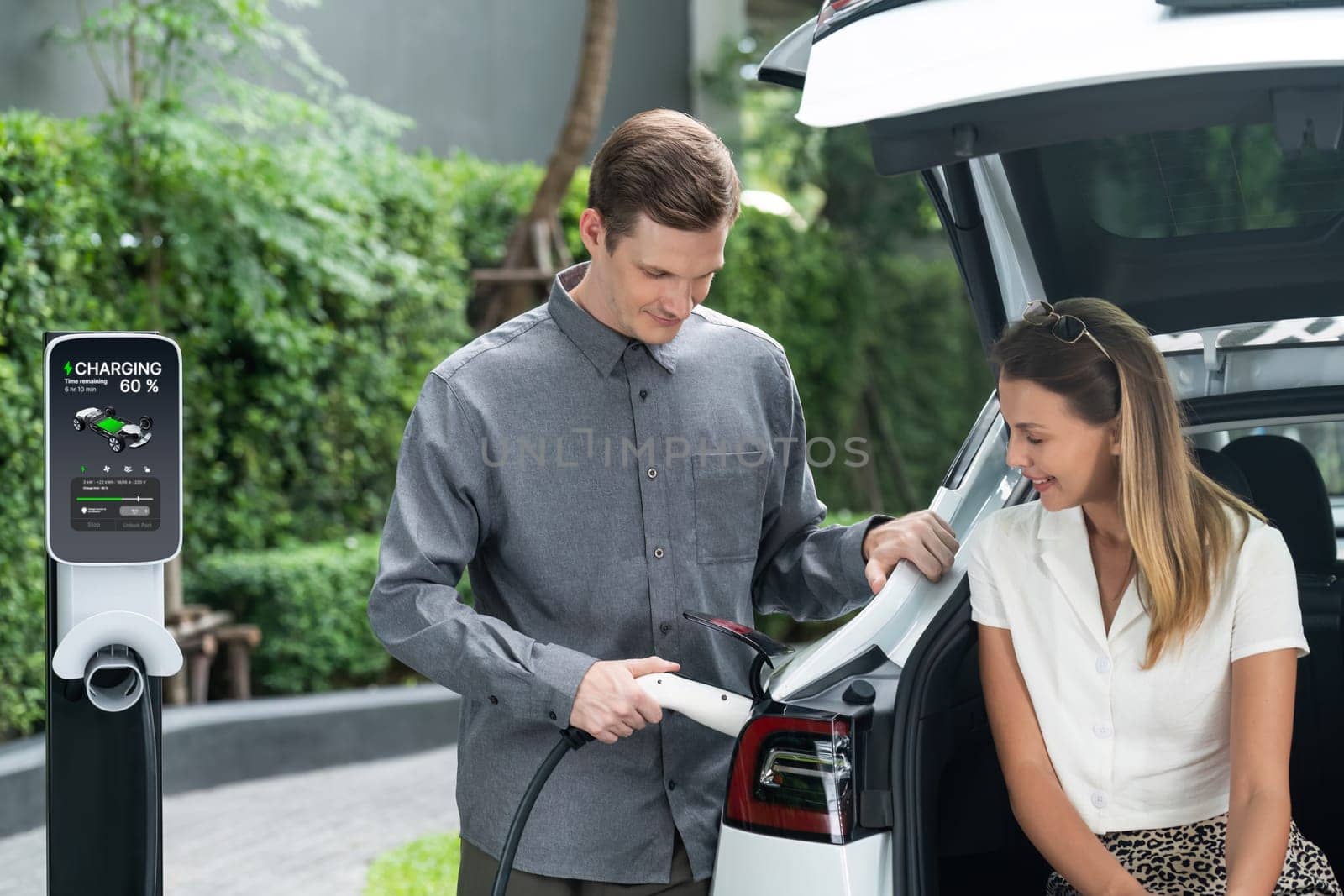 Young couple travel with EV electric car charging in green sustainable city outdoor garden in summer shows urban sustainability lifestyle by green clean rechargeable energy of electric vehicle innards