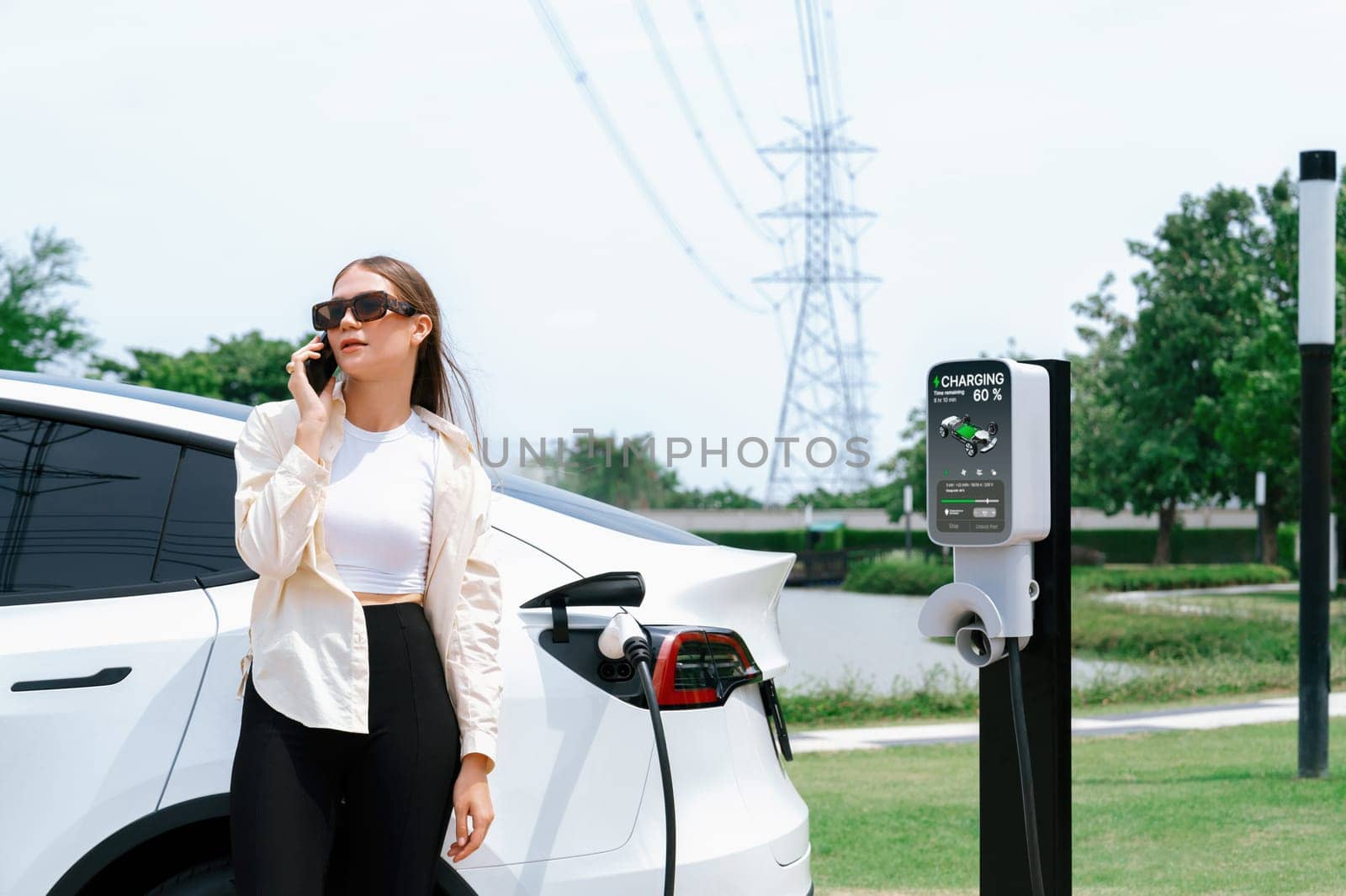 Young woman recharging EV car battery while talk on phone at charging station connected to electrical power grid tower facility as electrical industry for eco friendly vehicle utilization. Expedient