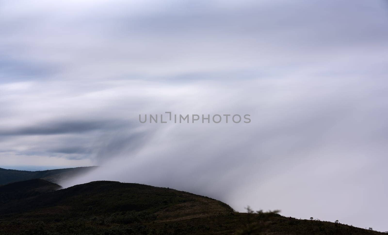 A long exposure shot captures the mist attempting to overcome the mountain peak, only to be stopped by strong winds from the opposite direction. The resulting effect is a stunning column of clouds reaching up towards the sky, creating a breathtaking landscape that is perfect for your next project.