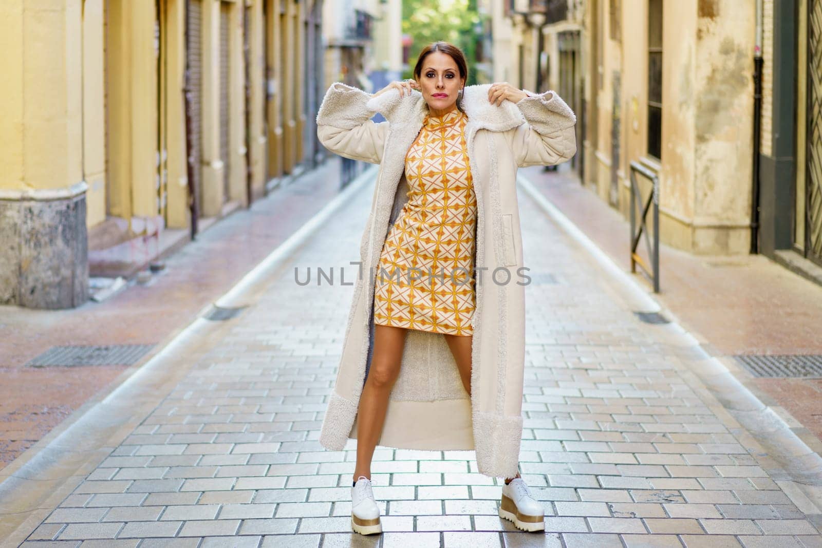 Confident young female wearing white coat and sneakers standing on paved street while touching neck of stylish outfit looking at camera against building