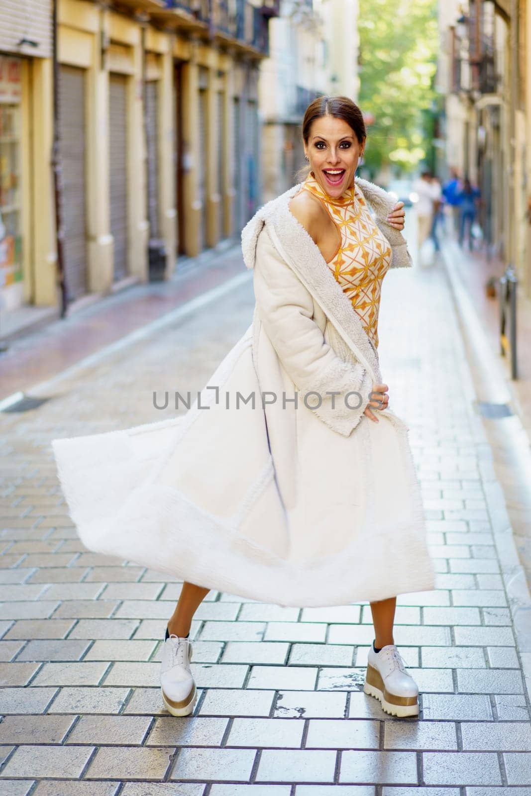 Smiling elegant young female wearing stylish coat and shoes standing on paved street against blurred old buildings and looking at camera