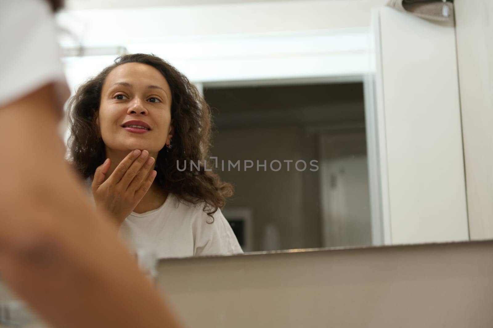 Smiling brunette, young pretty woman touching her face while admiring herself in the mirror of a home bathroom