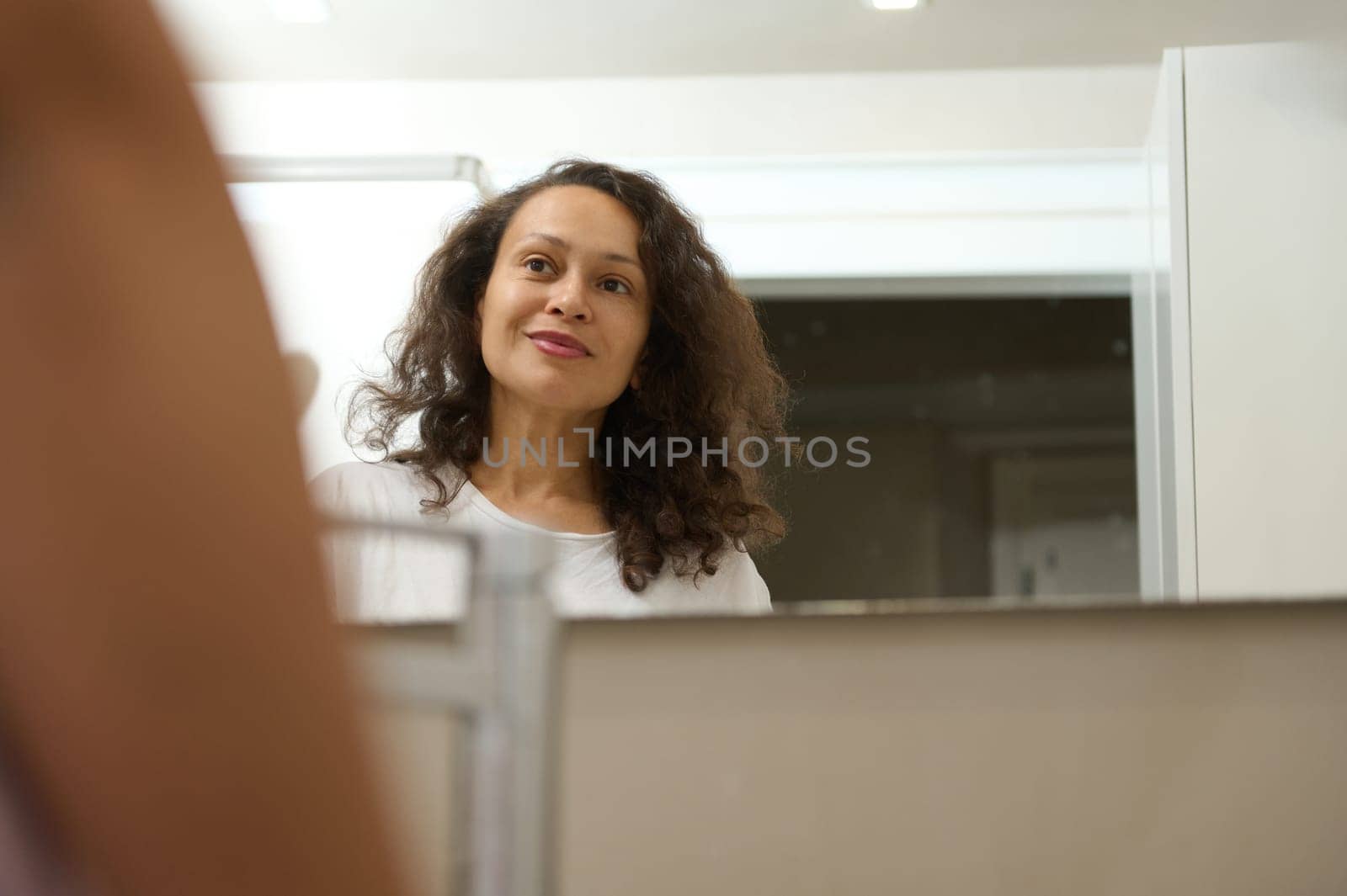 Reflection in the bathroom mirror of a multi ethnic smiling happy woman admiring her appearance. Home spa and beauty by artgf