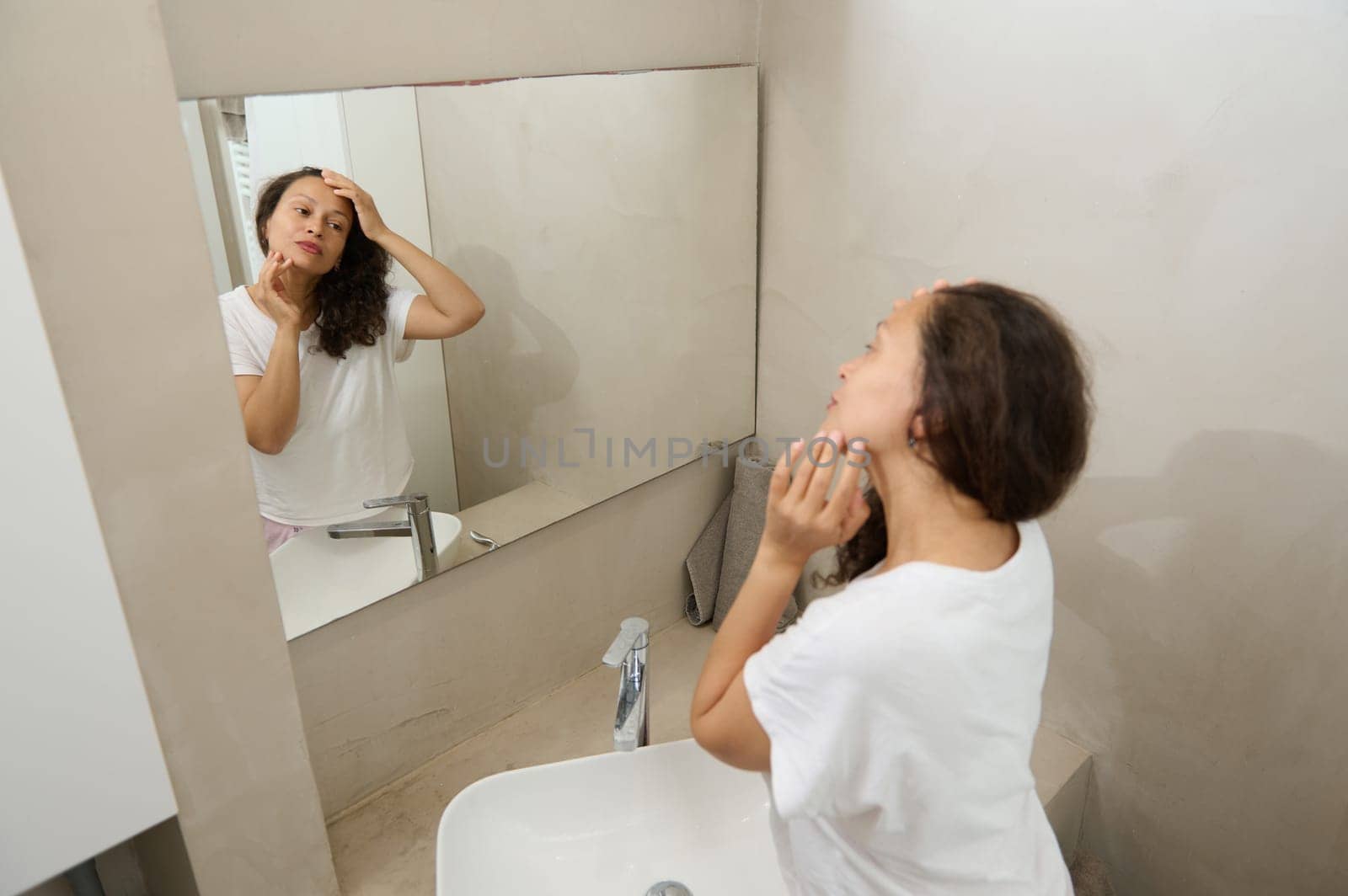 Happy young woman touching her face, examining flaws, skin problems and wrinkles, standing in front of a bathroom mirror by artgf