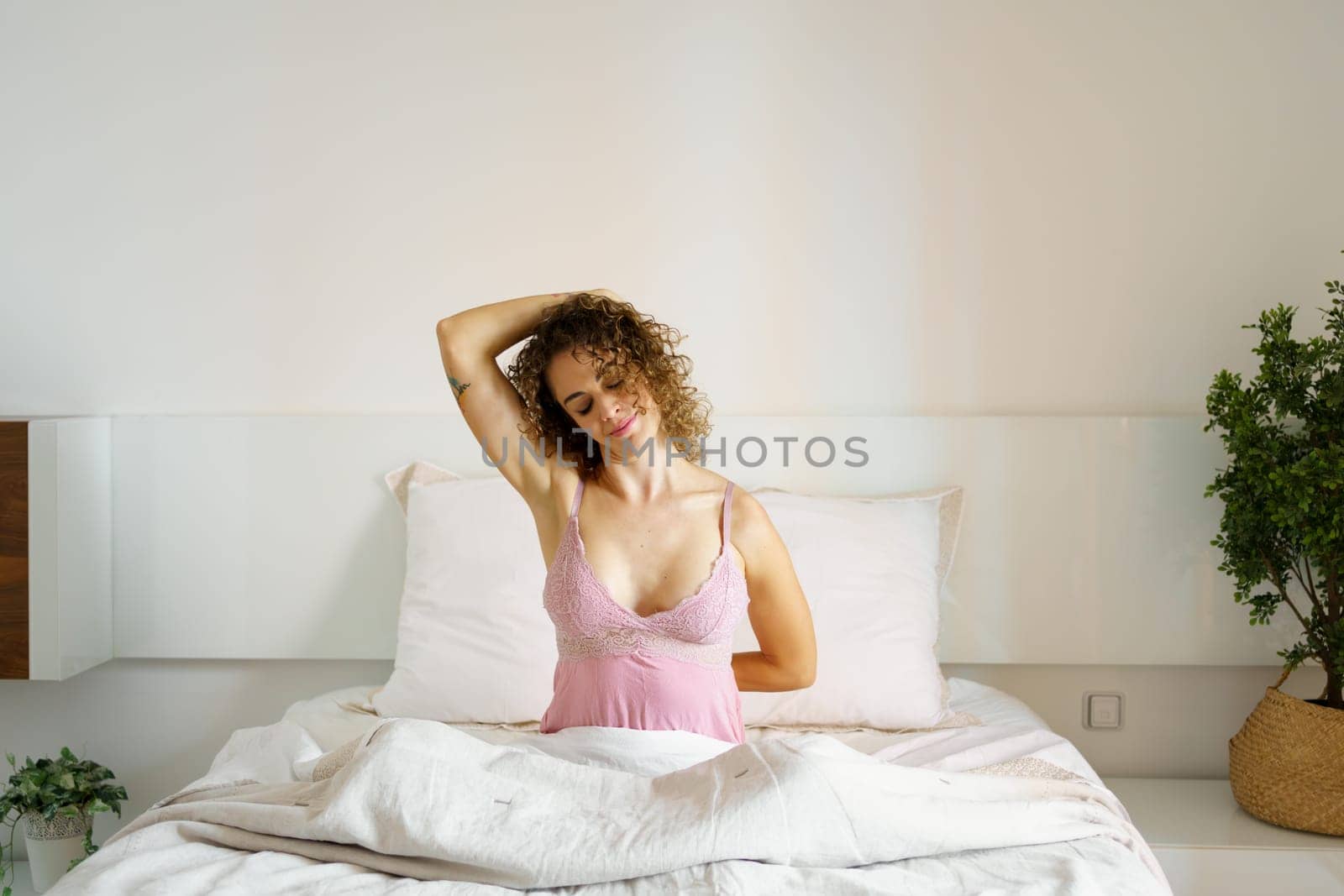 Smiling adult curly haired female with eyes closed while sitting doing stretching exercise, with hand on tilted head and with folded arm on back on cozy bed in daylight