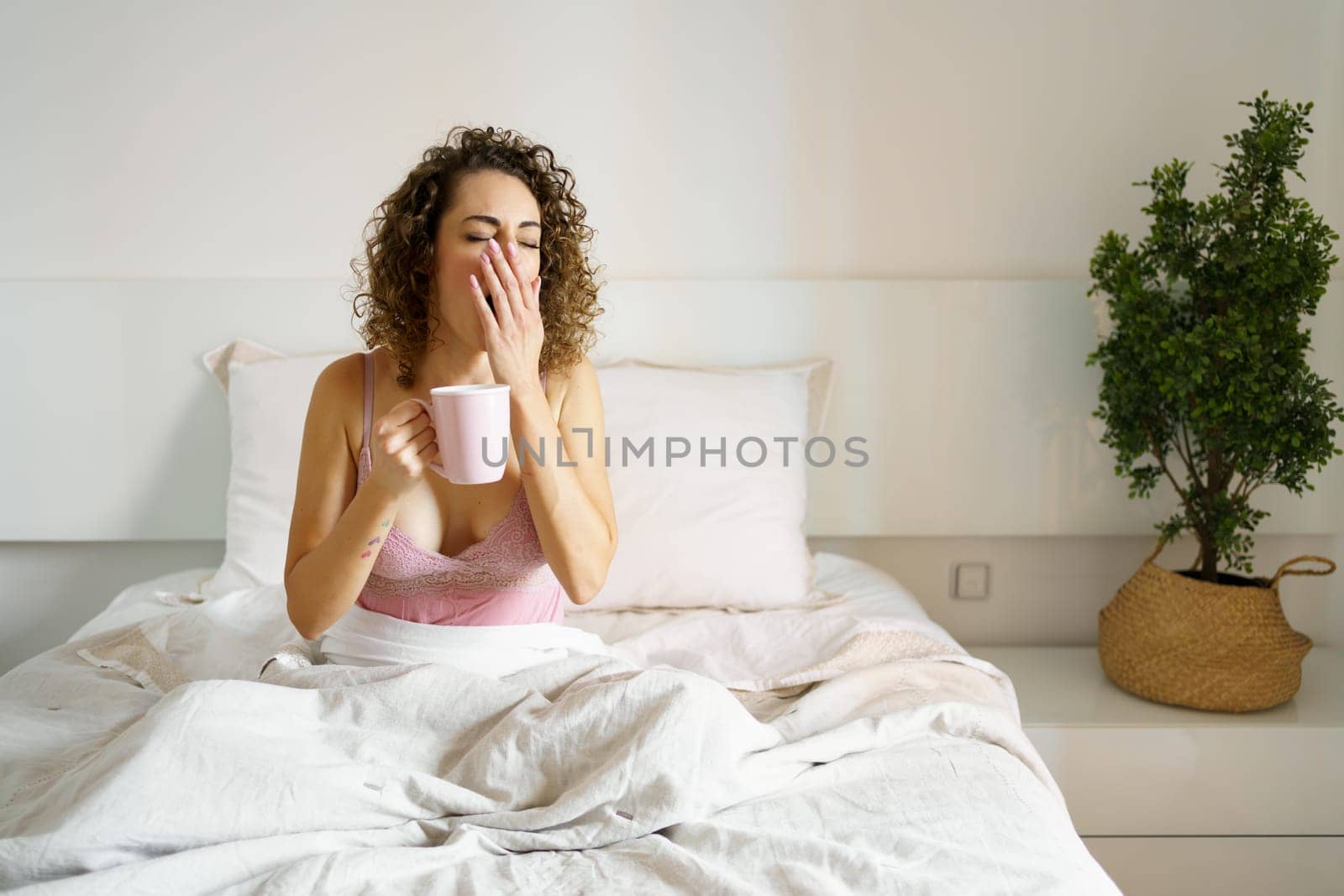 Young woman in nightwear yawning while holding coffee mug in bed during morning