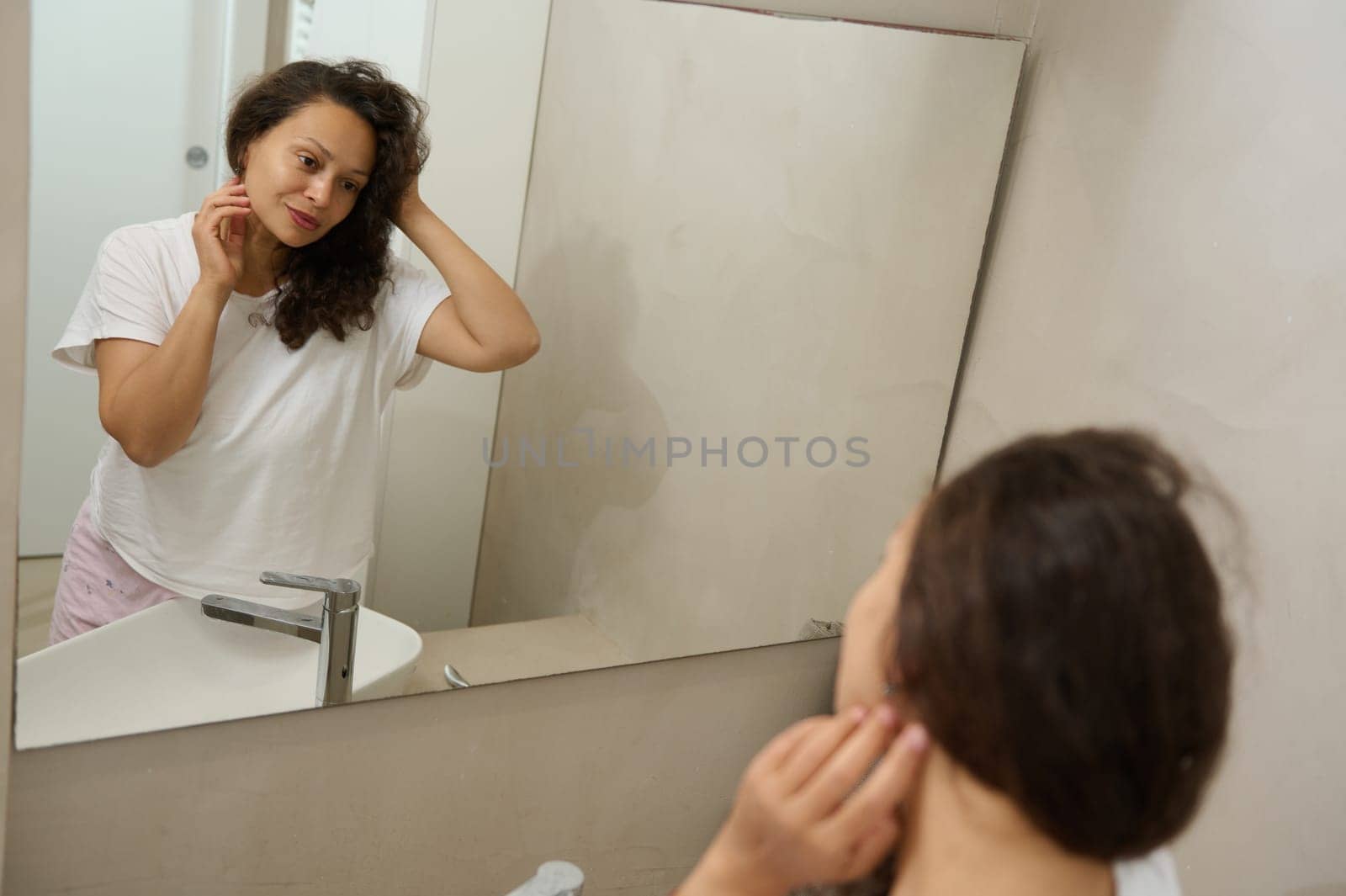Attractive multiracial young woman looking in the bathroom mirror and touching her face, enjoying her appearance. People. Healthy lifestyle. Home spa. Beauty, hygiene and skin care concept