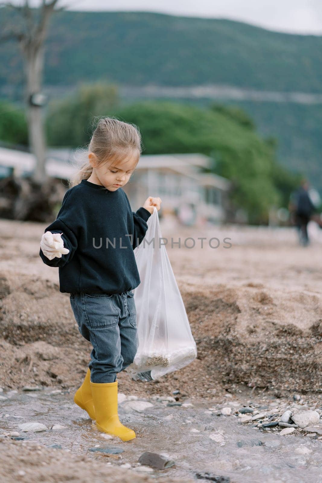 Little girl in rubber boots and gloves with a bag walks through shallow water looking for garbage by Nadtochiy