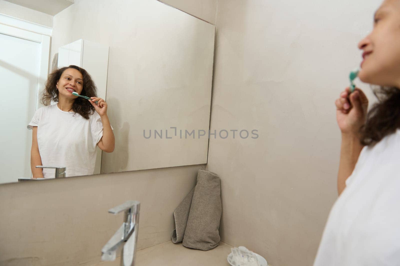 Reflection in the bathroom mirror of a happy pretty young woman in pajamas, brushing teeth in the morning, standing by a ceramic sink in the home bathroom