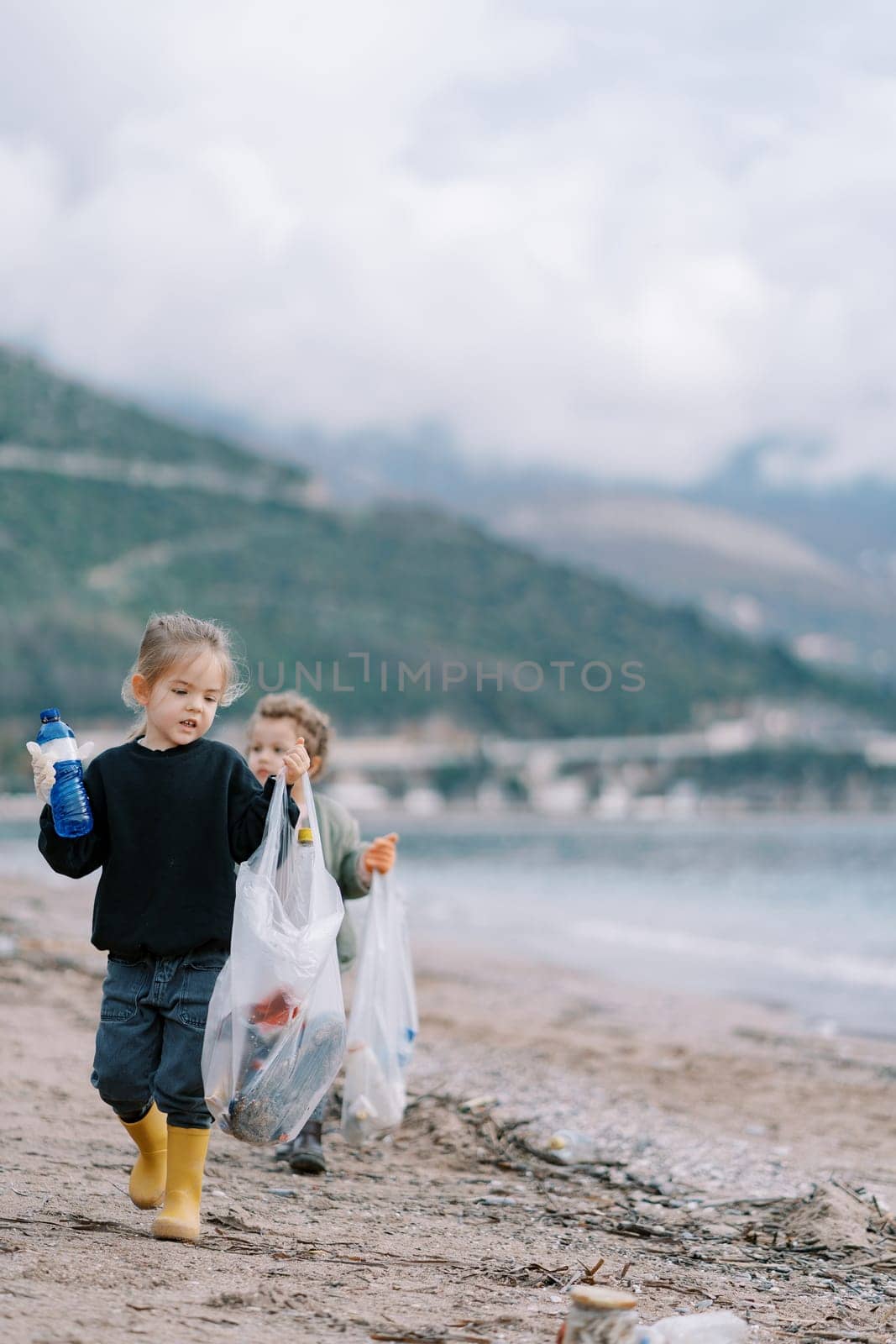 Little girls collect plastic bottles into bags on the sandy beach. High quality photo