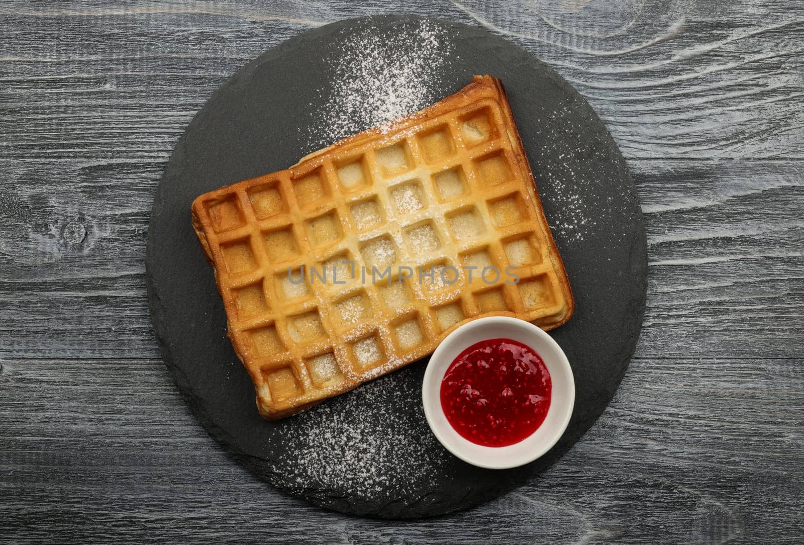Close up big sweet traditional American or Belgian Brussels waffle with powder sugar and raspberry jam served on black slate board on table, elevated top view, directly above