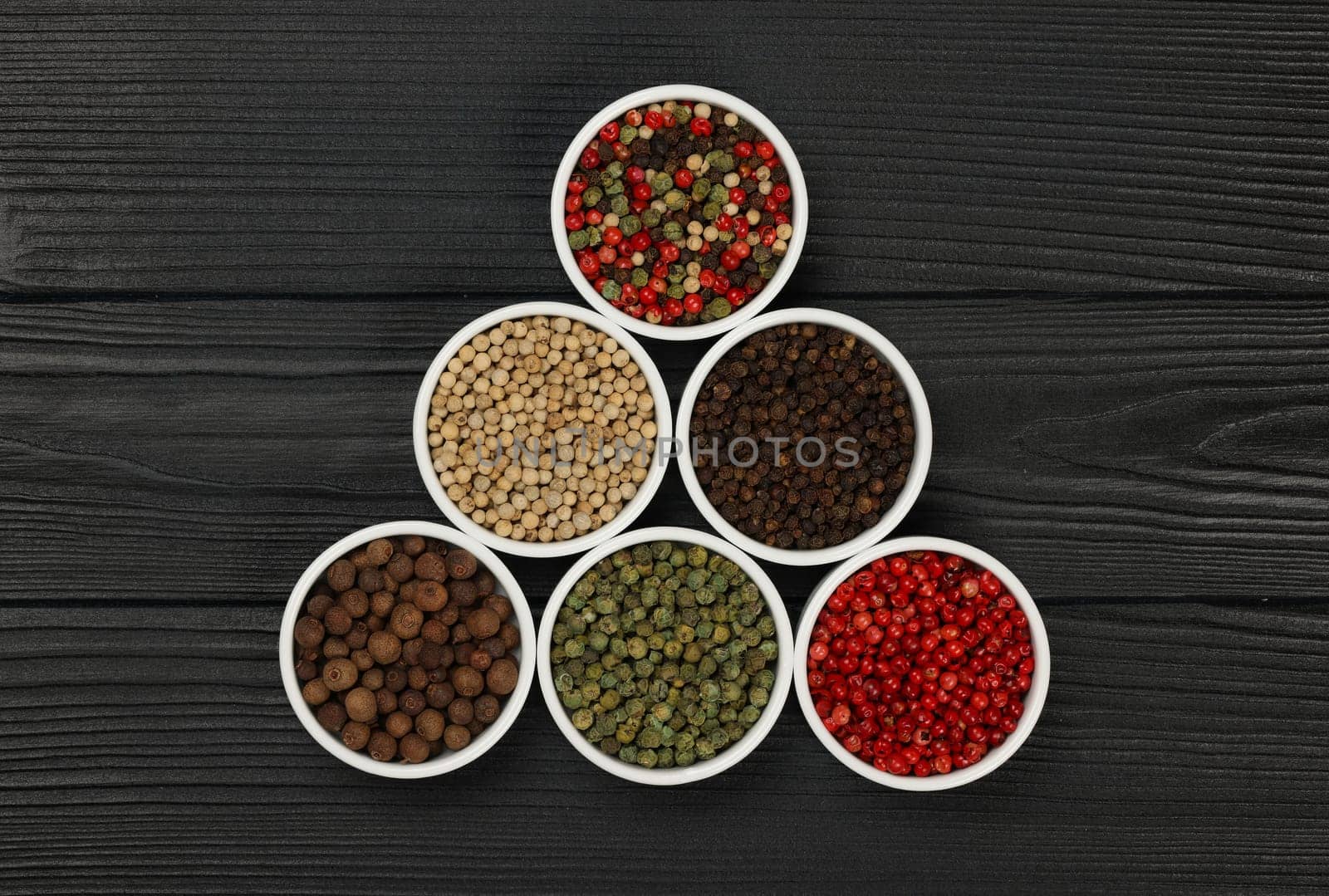 Triangle of assorted peppercorns in white ceramic bowls over black wooden table background with copy space, elevated top view, directly above