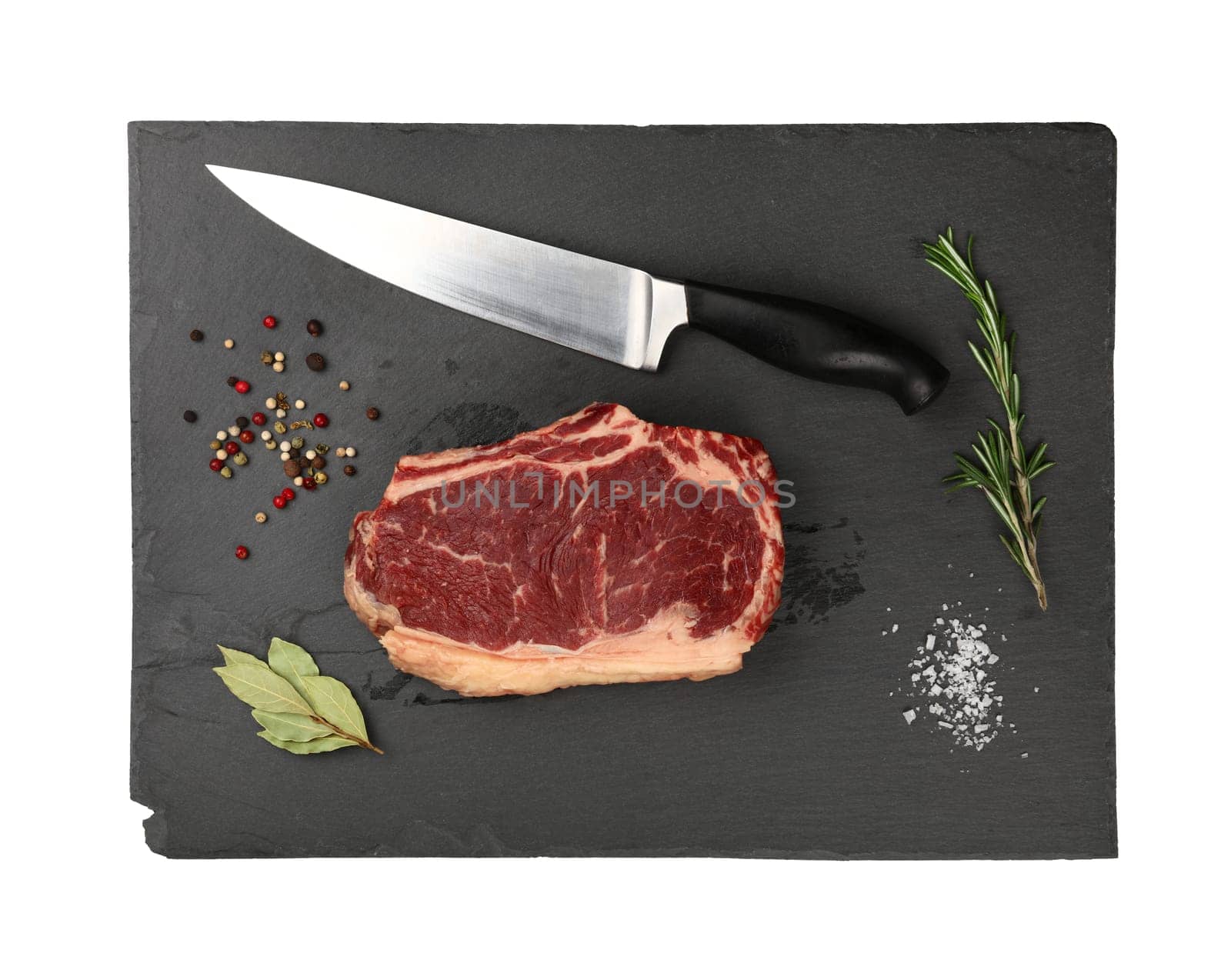 Close up one marbled raw ribeye beef steak with rib bone on black slate cutting board with knife and spices to season, isolated on white background, elevated top view, directly above