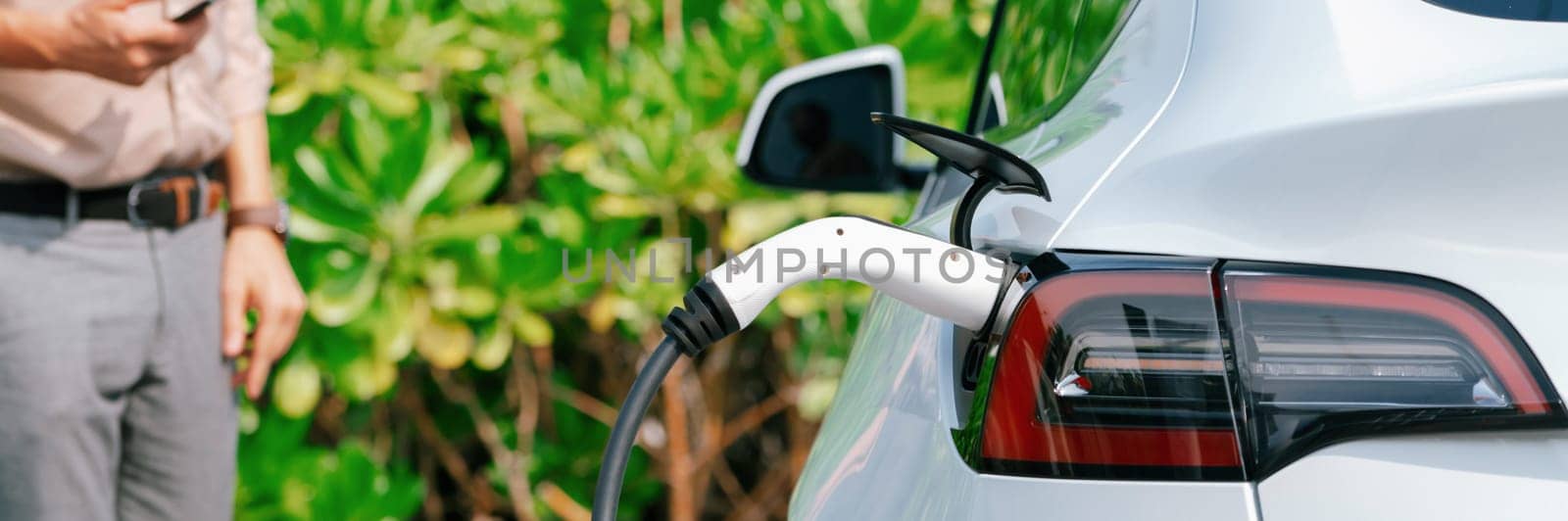 Young man use smartphone to pay for electricity at public EV car charging station green city park. Modern environmental and sustainable urban lifestyle with EV vehicle. Panorama Expedient