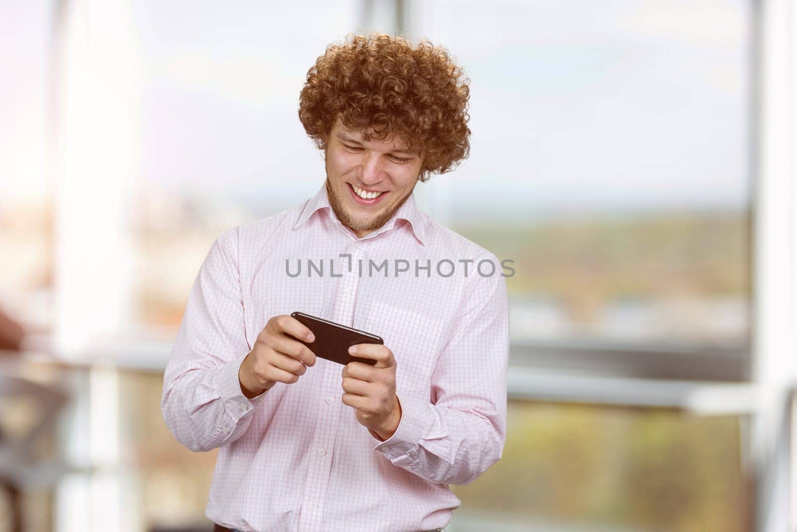 Excited happy young caucasian male gamer playing using smartphone. Guy playing mobile videogame app or watching digital video stream holding cell phone indoors.