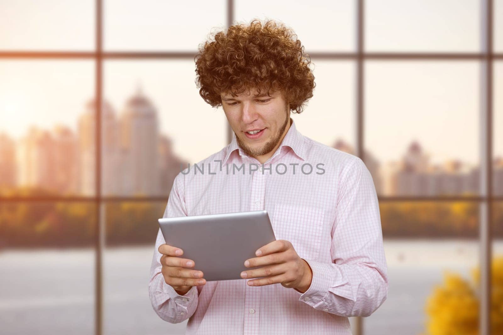 Portrait of a young caucasian man with curly hair playing a video game on the tablet pc device. Indoor window in the background.