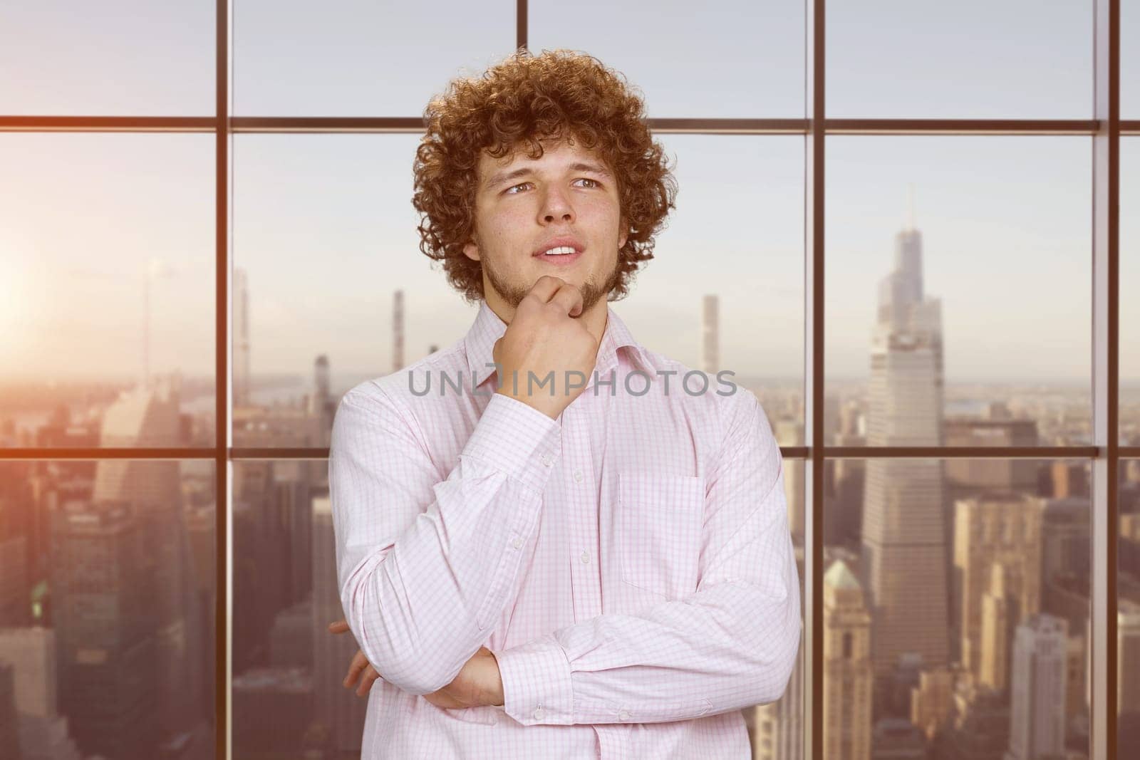 Portrait of a pensive young businessman with curly hair touching his chin. Having an idea. Indoor window with cityscape view in the background.