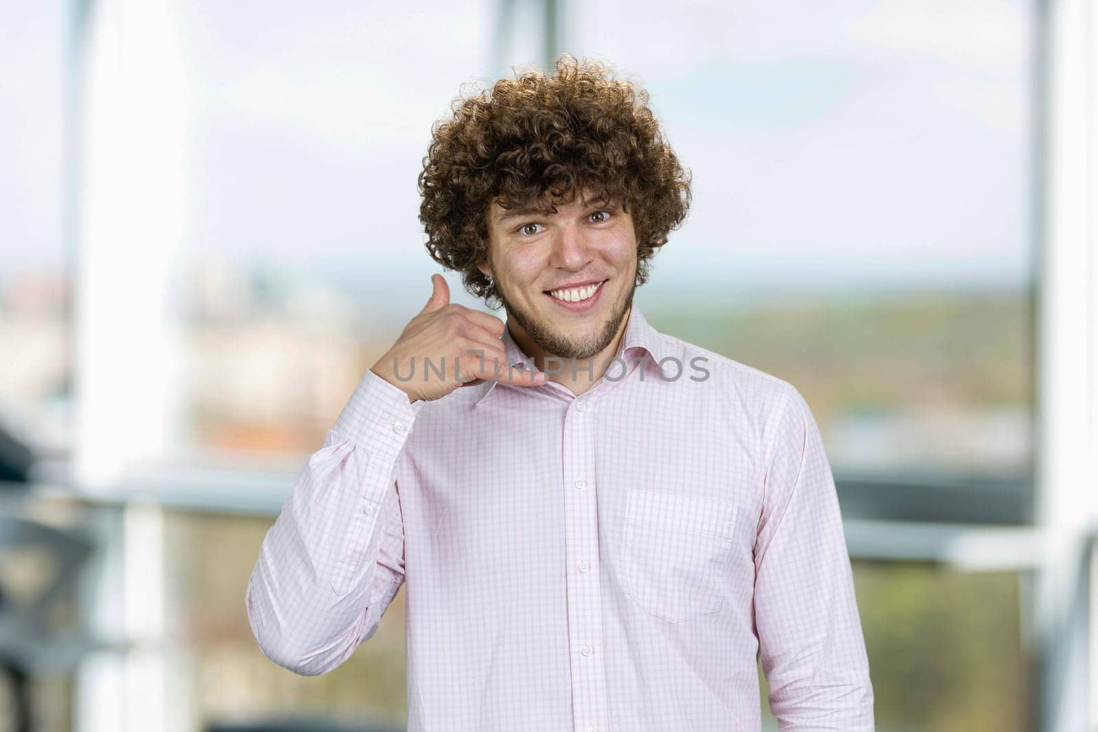 Handsome young man with curly hair shows call me gesture. Indoor window in the background.