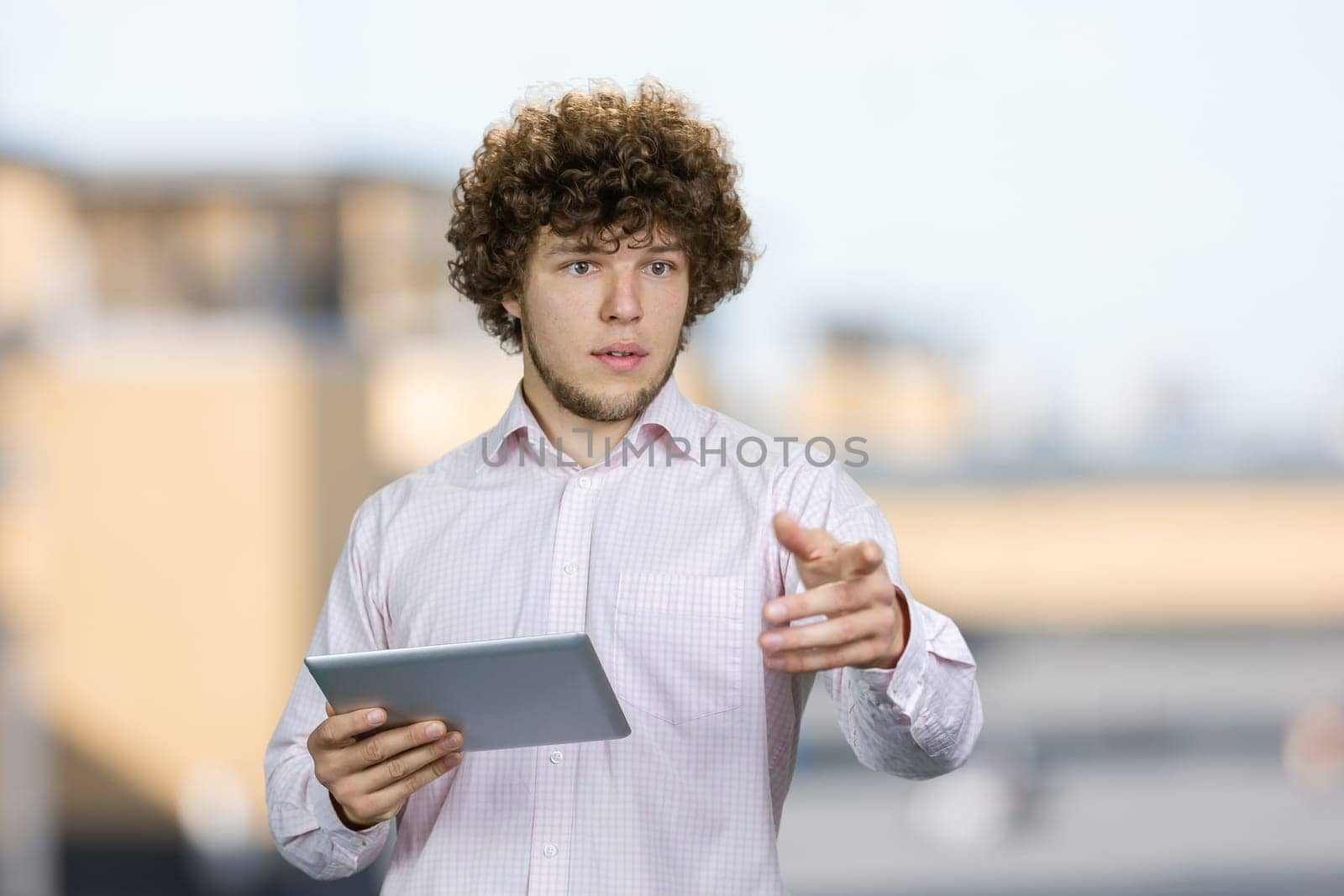 Portrait of a young man with curly hair holding a tablet pc and pointing at something. Blurred urban background.